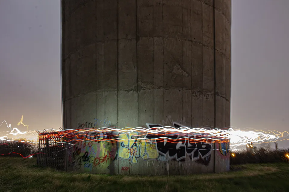 Ribbons of white and orange light drawn around the graffitied base of a concrete tower at dusk